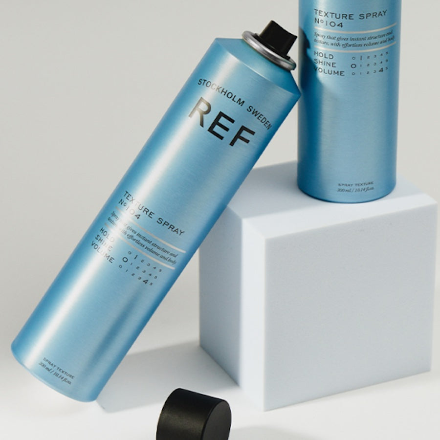 Genuine REF product photo (authorized distributor asset pack) — styled shot of two Texture Spray N°104 cans leaning against a white block, editorial/lifestyle framing.